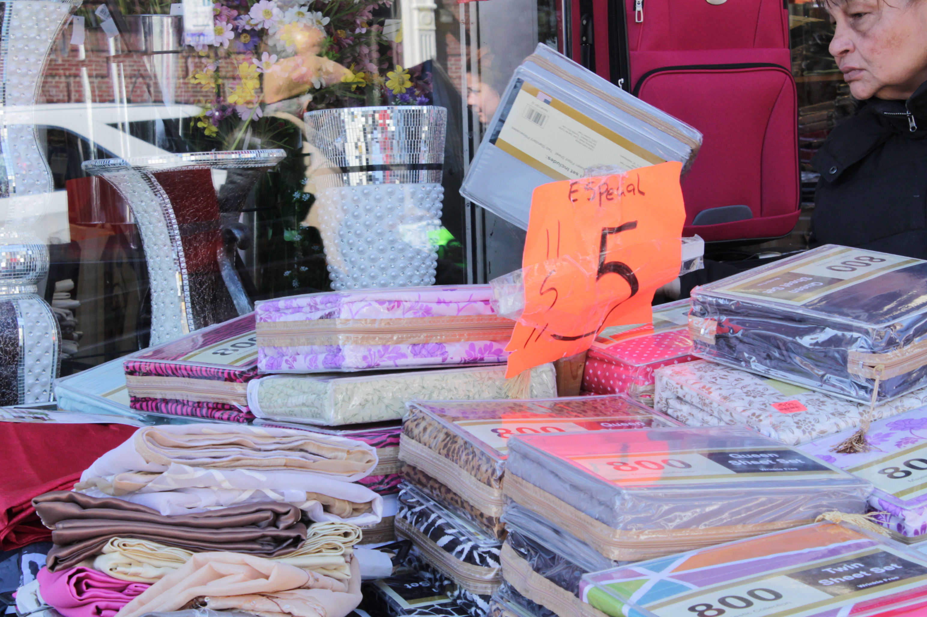 Photograph of a table with many different types of fabric for sale. There is a bright pink paper sign in the center of the table that says Sale in Spanish